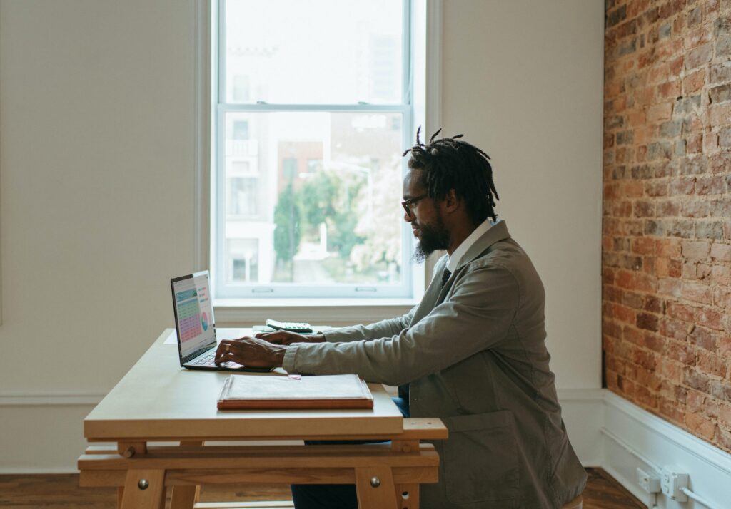 A photo of a man in a suit sitting at a desk with a laptop and papers around him. Given the unconventional nature of his office, it is assumed he works remotely.